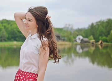 A young woman with long brown hair, smiling and standing by a lake.