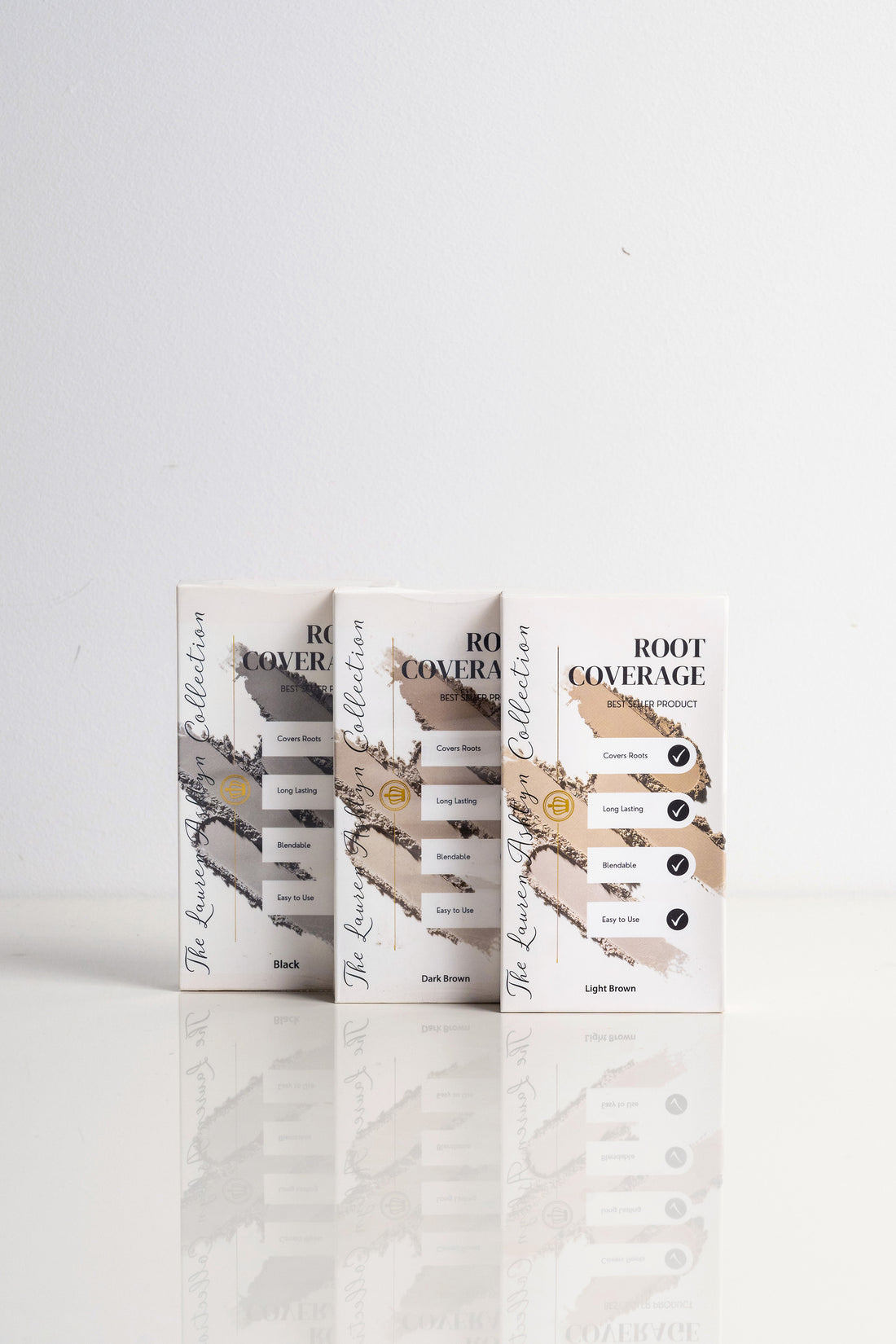 Three boxes of Lauren Ashtyn root coverage powder leaning upright side-by-side in shades of black, dark brown, and light brown.