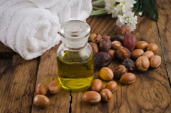 A glass bottle of argan oil placed on a wooden surface, surrounded by argan nuts and seeds. A white rolled towel and fresh flowers are set in the background, creating a natural and spa-like ambiance.