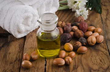 A glass bottle of argan oil placed on a wooden surface, surrounded by argan nuts and seeds. A white rolled towel and fresh flowers are set in the background, creating a natural and spa-like ambiance.