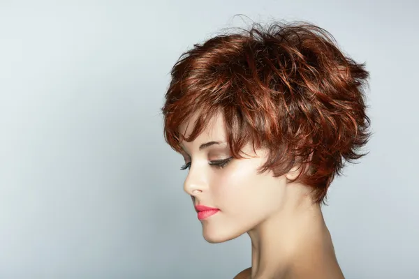 A woman with a short, textured auburn hairstyle, showcasing soft waves and layers. She is gazing downward with subtle makeup, including a bold pink lip, against a neutral gray background. 