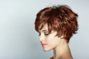 A woman with a short, textured auburn hairstyle, showcasing soft waves and layers. She is gazing downward with subtle makeup, including a bold pink lip, against a neutral gray background. 