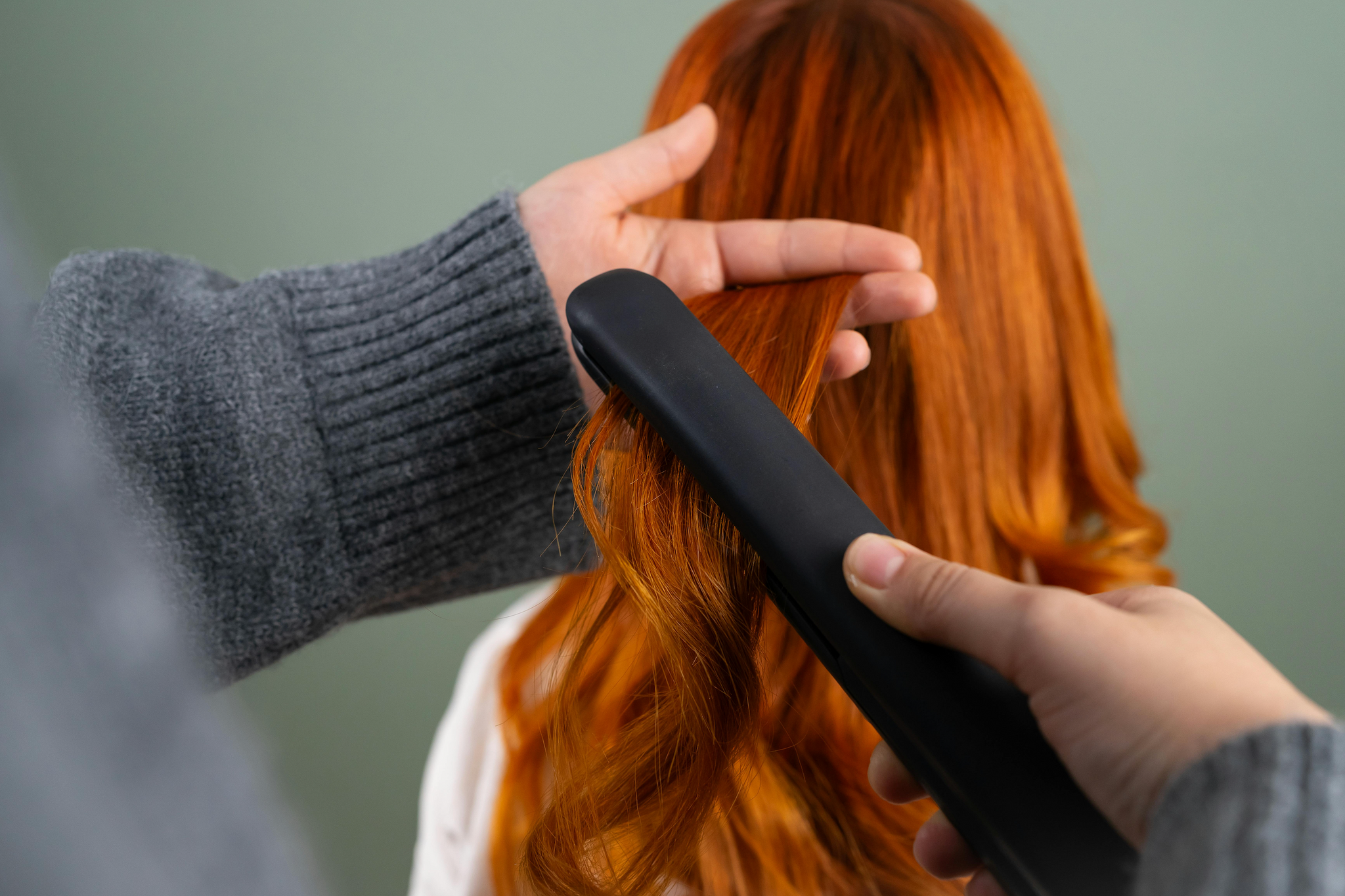 A person adding volume to a woman’s bright auburn hair using a flat iron.
