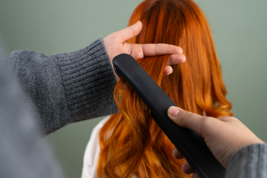 A person adding volume to a woman’s bright auburn hair using a flat iron.