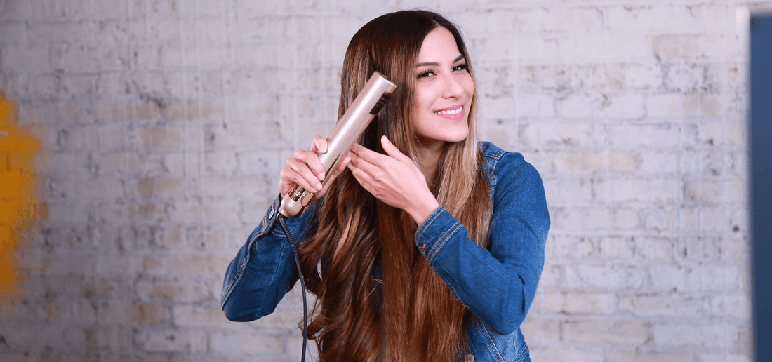 A brunette woman with long hair styles her hair with a TYME Iron Pro against a white brick back drop. 