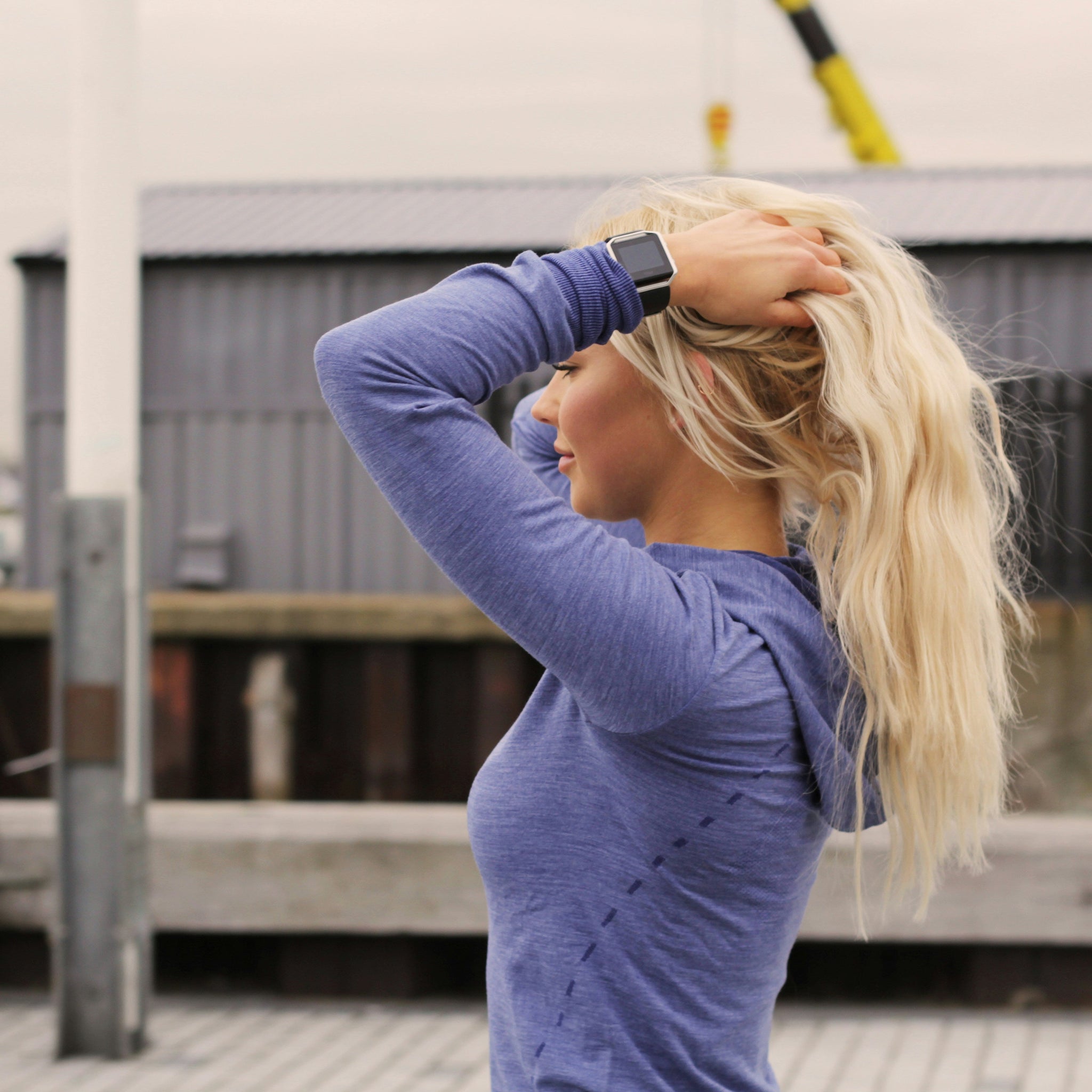 Woman in a blue long-sleeve athletic top holding her blonde hair up after a workout, standing outdoors near a dock — representing post-gym hair refresh.