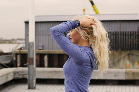 Woman in a blue long-sleeve athletic top holding her blonde hair up after a workout, standing outdoors near a dock — representing post-gym hair refresh.