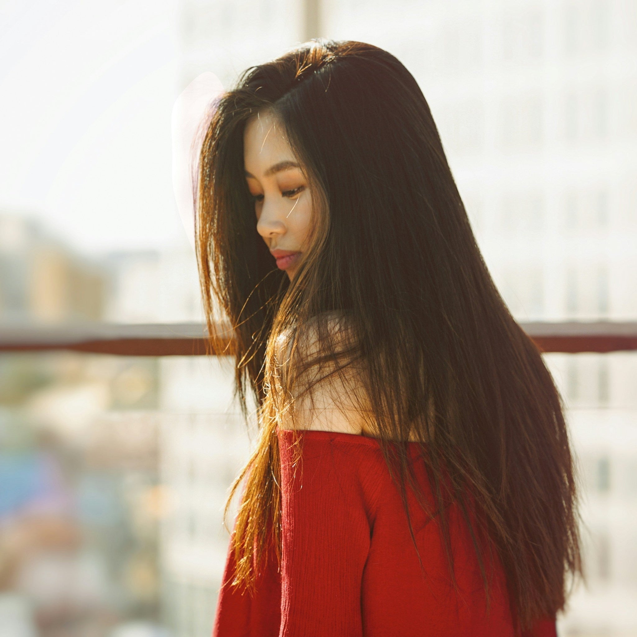 Woman with long, smooth hair standing near a window in sunlight, wearing an off-the-shoulder red sweater — showcasing sleek, healthy hair styled with an eco-friendly flat iron.