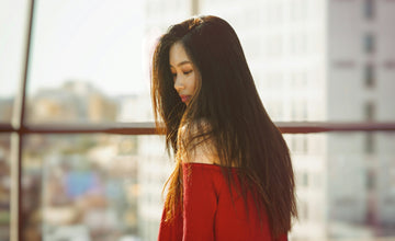 Woman with long, smooth hair standing near a window in sunlight, wearing an off-the-shoulder red sweater — showcasing sleek, healthy hair styled with an eco-friendly flat iron.