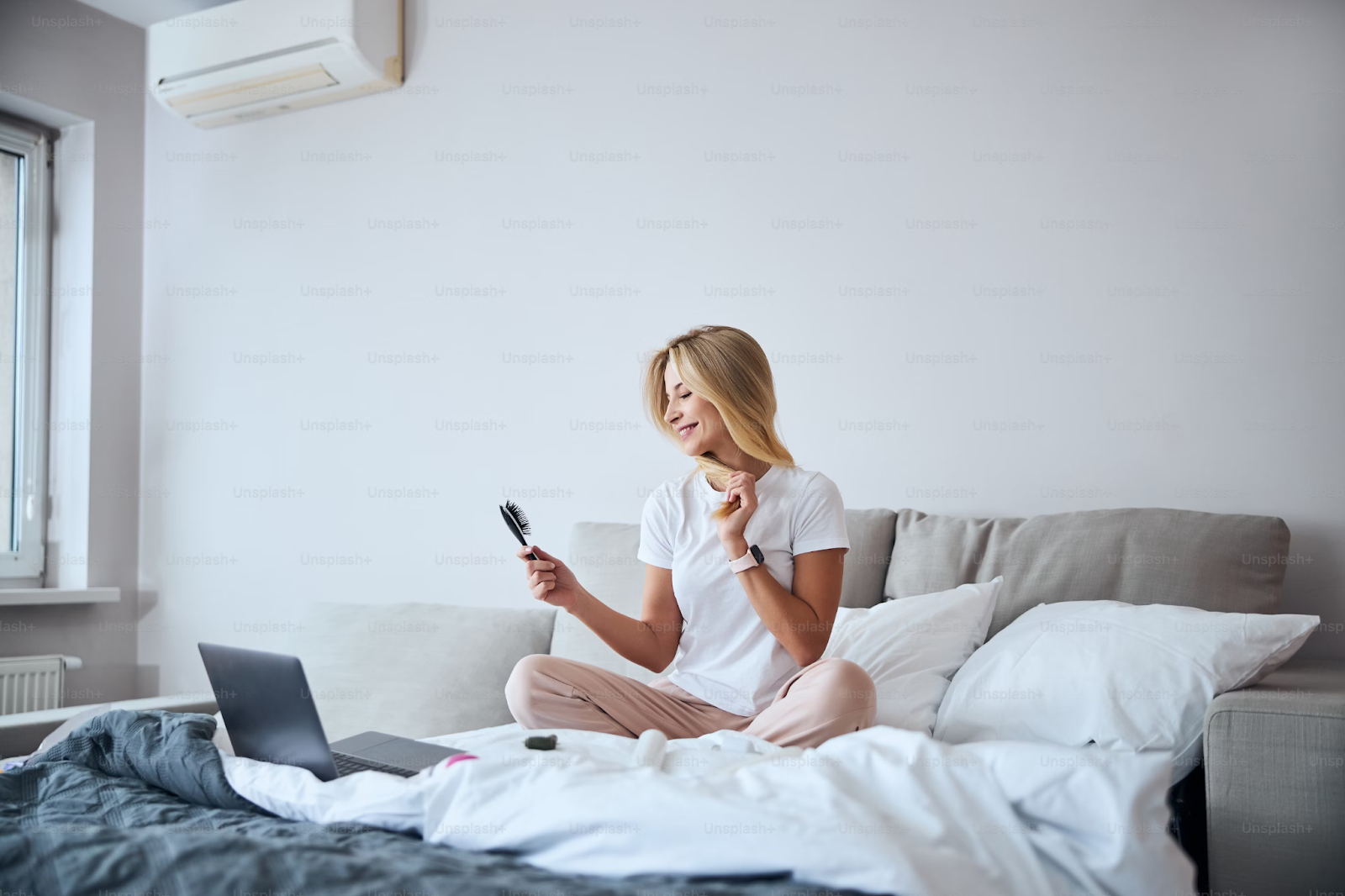 A smiling young woman sat on her bed in front of a laptop, brushing her hair.