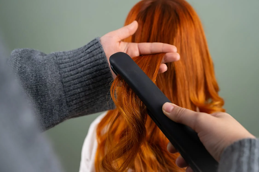 Hands using a flat iron to style long red hair into loose curls.