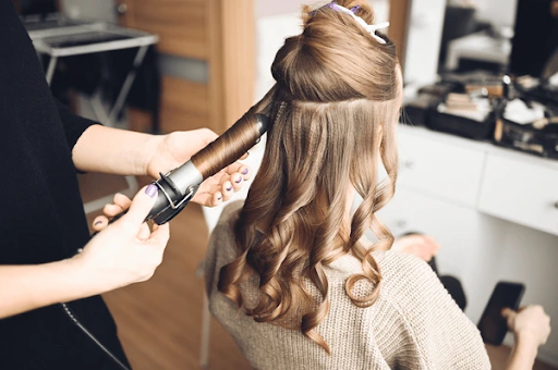 Person using a curling iron to style long, sectioned brown hair into soft curls in a salon setting, with a vanity mirror and hair tools in the background.