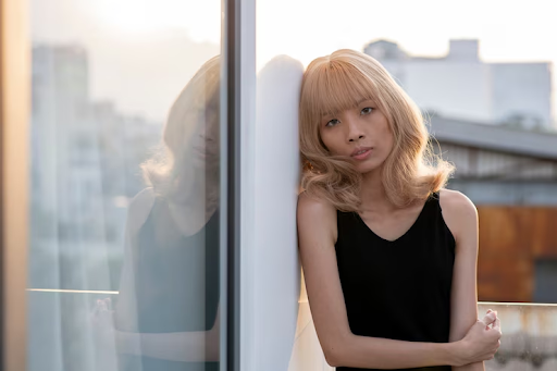 A fair-skinned female with blonde wavy hair and bangs leaning against a glass wall while looking at the camera