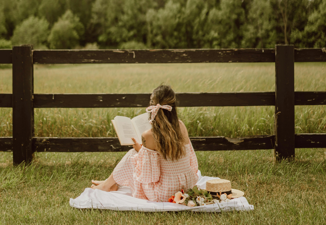 A woman sat on a picnic blanket in a field with a large bow in her hair.