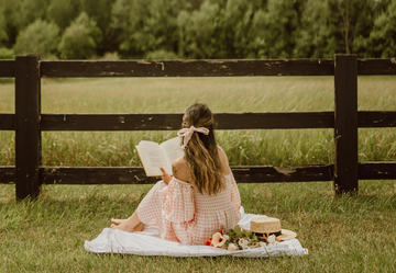 A woman sat on a picnic blanket in a field with a large bow in her hair.