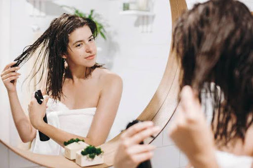 Young woman in front of a mirror, spraying product into damp, dark hair.