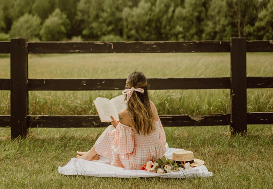 A woman sat on a picnic blanket in a field with a large bow in her hair.