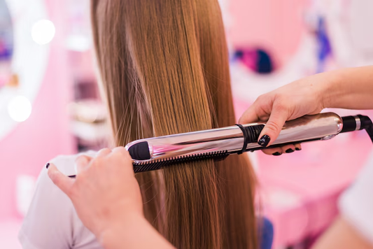 Hairstylist using a flat iron to straighten a client's long, light brown hair in a brightly lit salon with pink decor.