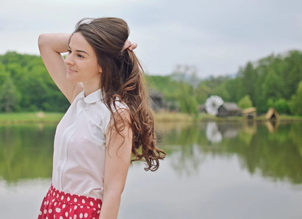 A young woman with long brown hair, smiling and standing by a lake.