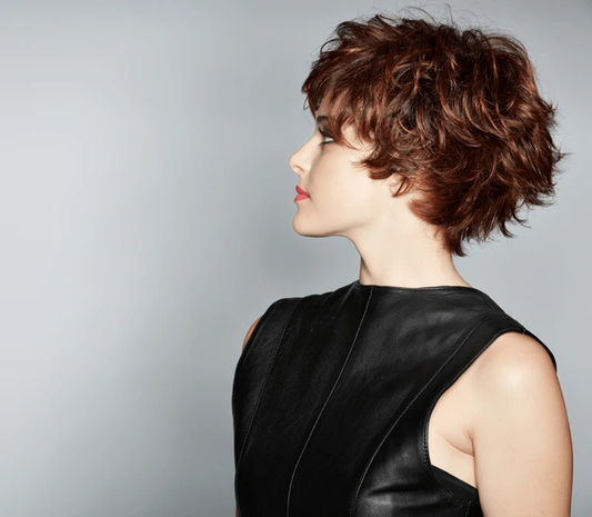 Woman with a short, tousled auburn pixie cut wearing a black sleeveless leather top, shown in profile against a light gray background.